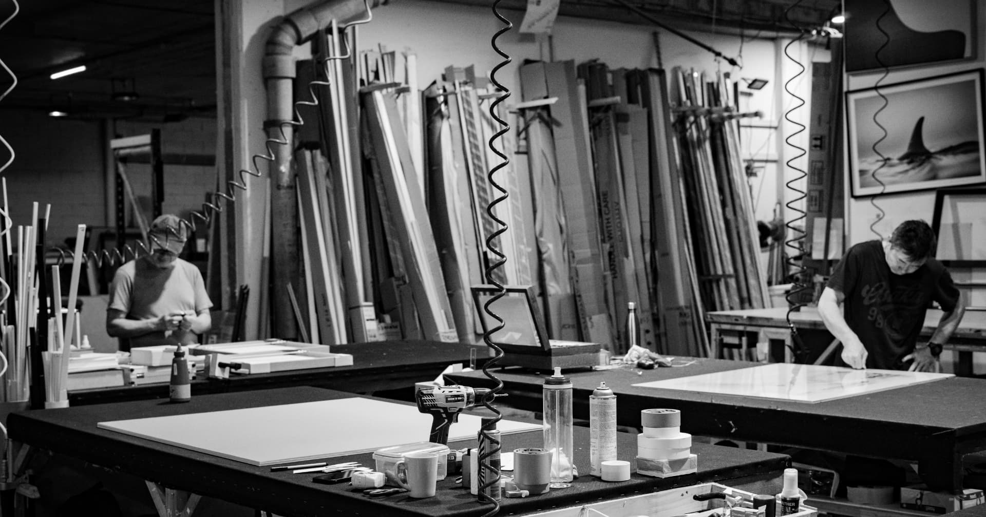 Two framers at their benches, moulding lengths racked on the wall behind them
