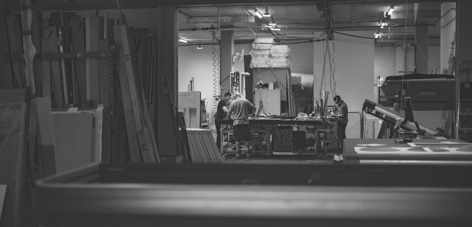 Looking through a doorway onto the workshop floor: four people at the bench, moulding lengths stacked along the wall