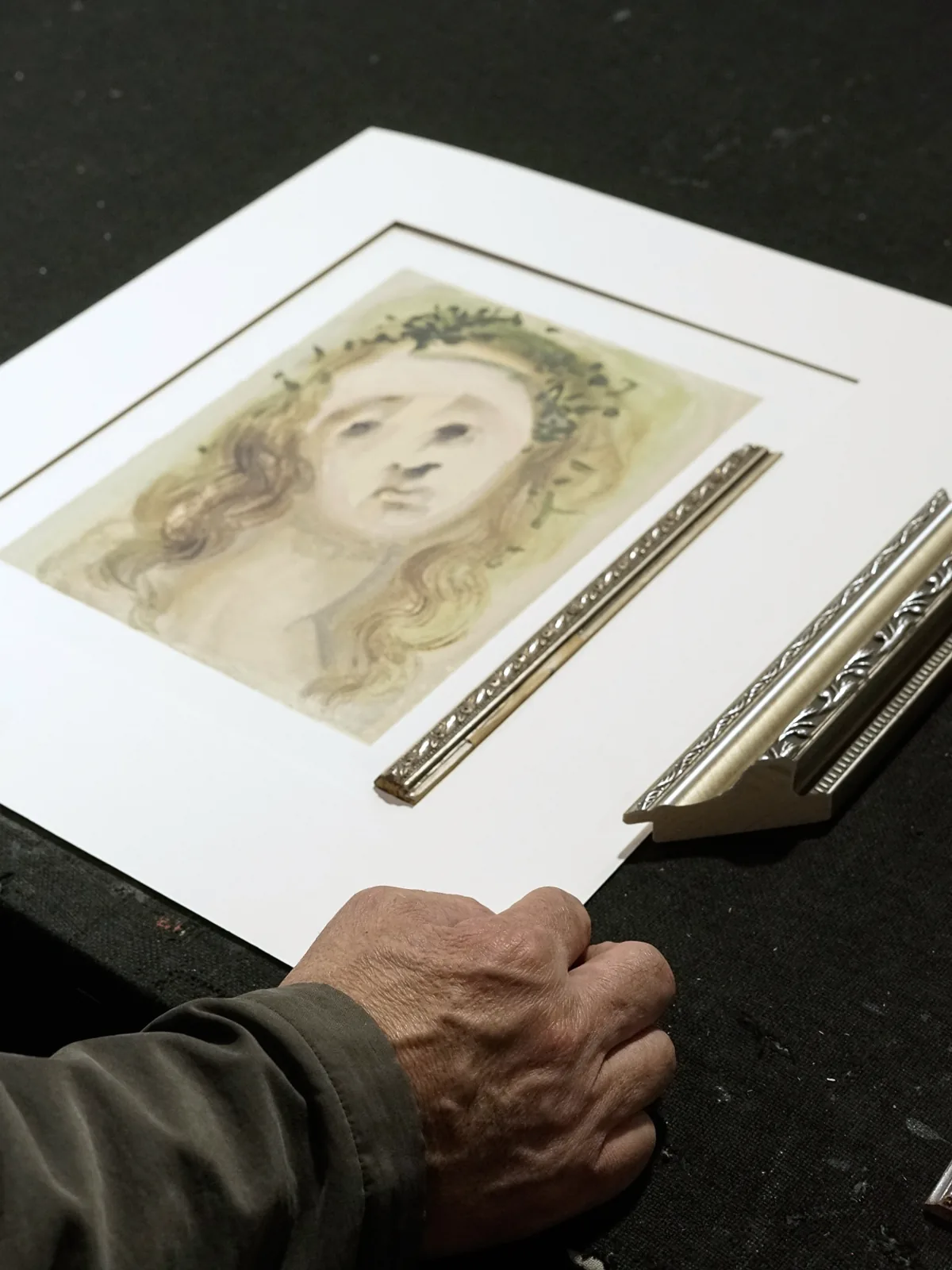 A framer's hand on a matted watercolour portrait, with two lengths of ornate silver moulding laid beside it