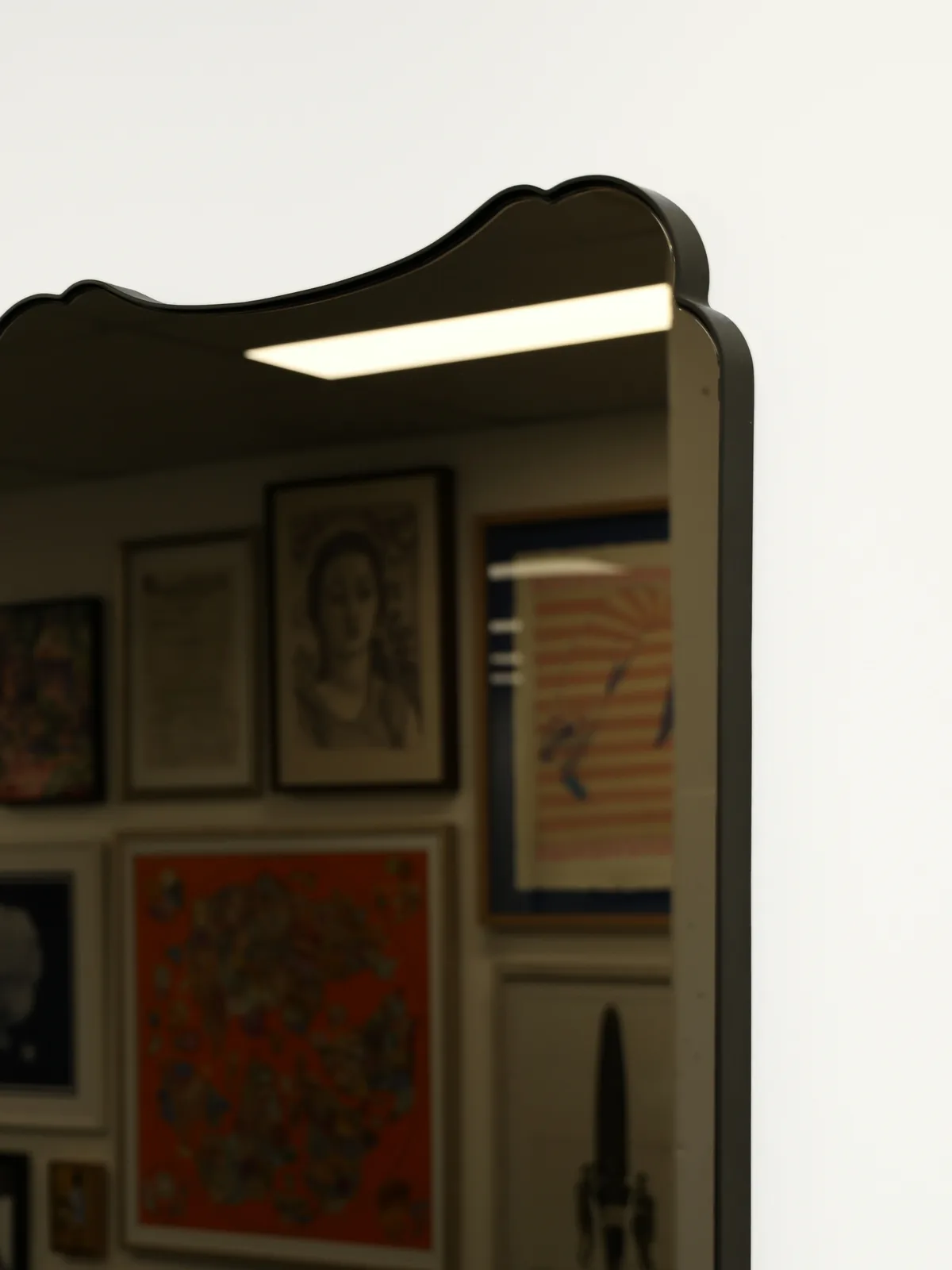 A black scalloped-top mirror on a white wall, reflecting a wall of framed art in the workshop