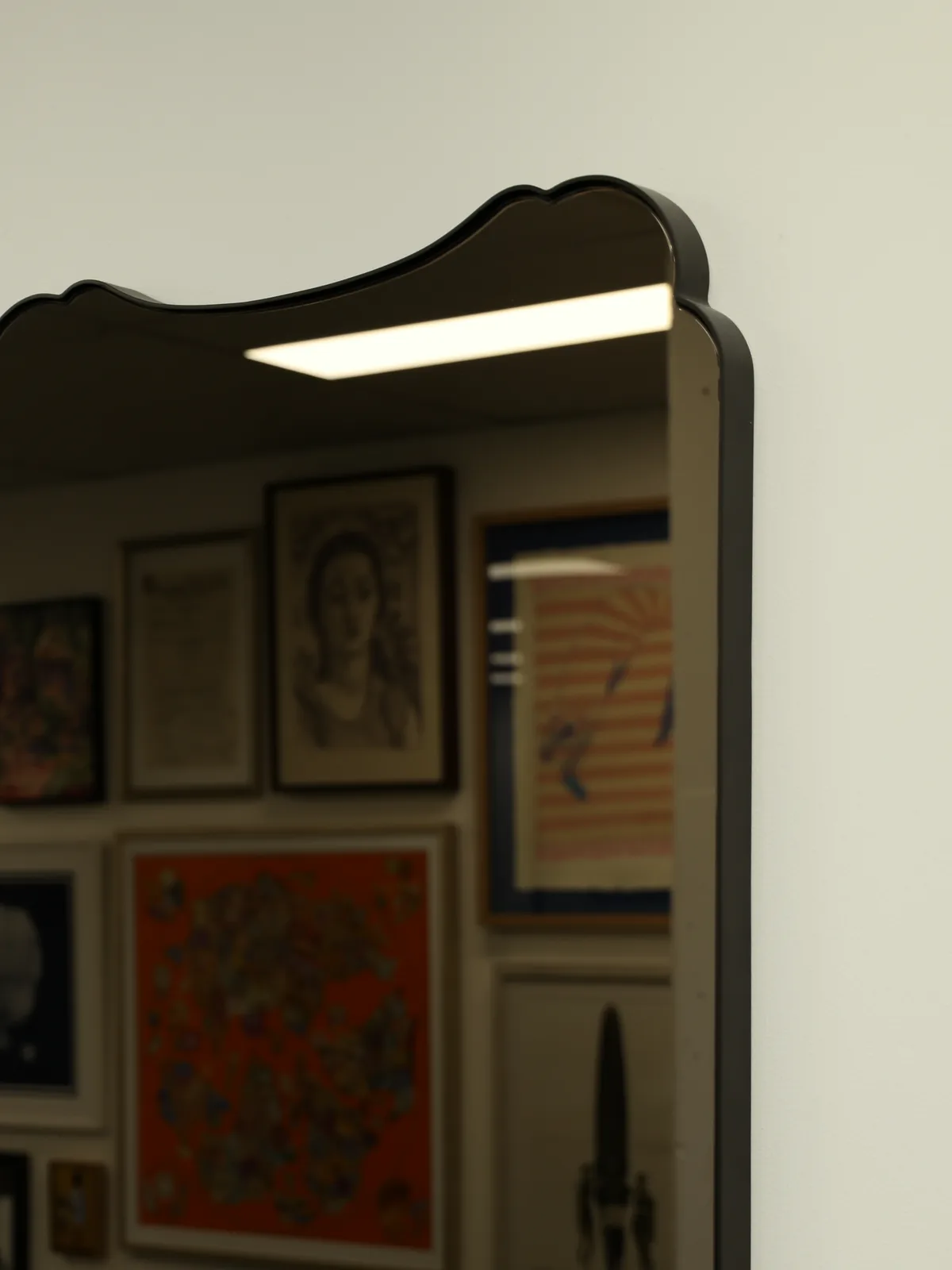 A black scalloped-top mirror on a white wall, reflecting a wall of framed art in the workshop