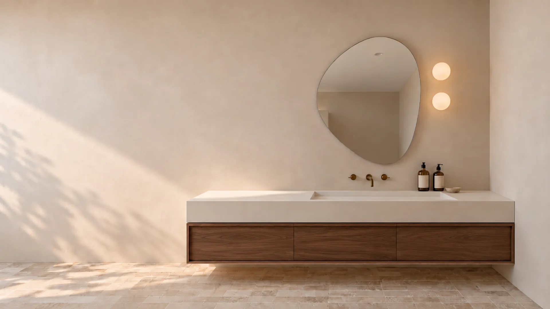 A frameless organic-shaped mirror above a floating walnut vanity with a stone top, on a warm plaster wall with two globe lights and afternoon leaf shadow