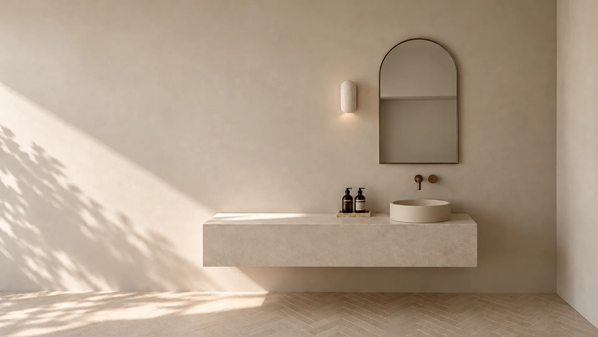A tall arched mirror in a slim brass frame above a floating stone vanity with a round basin, on a warm plaster wall in afternoon light and leaf shadow