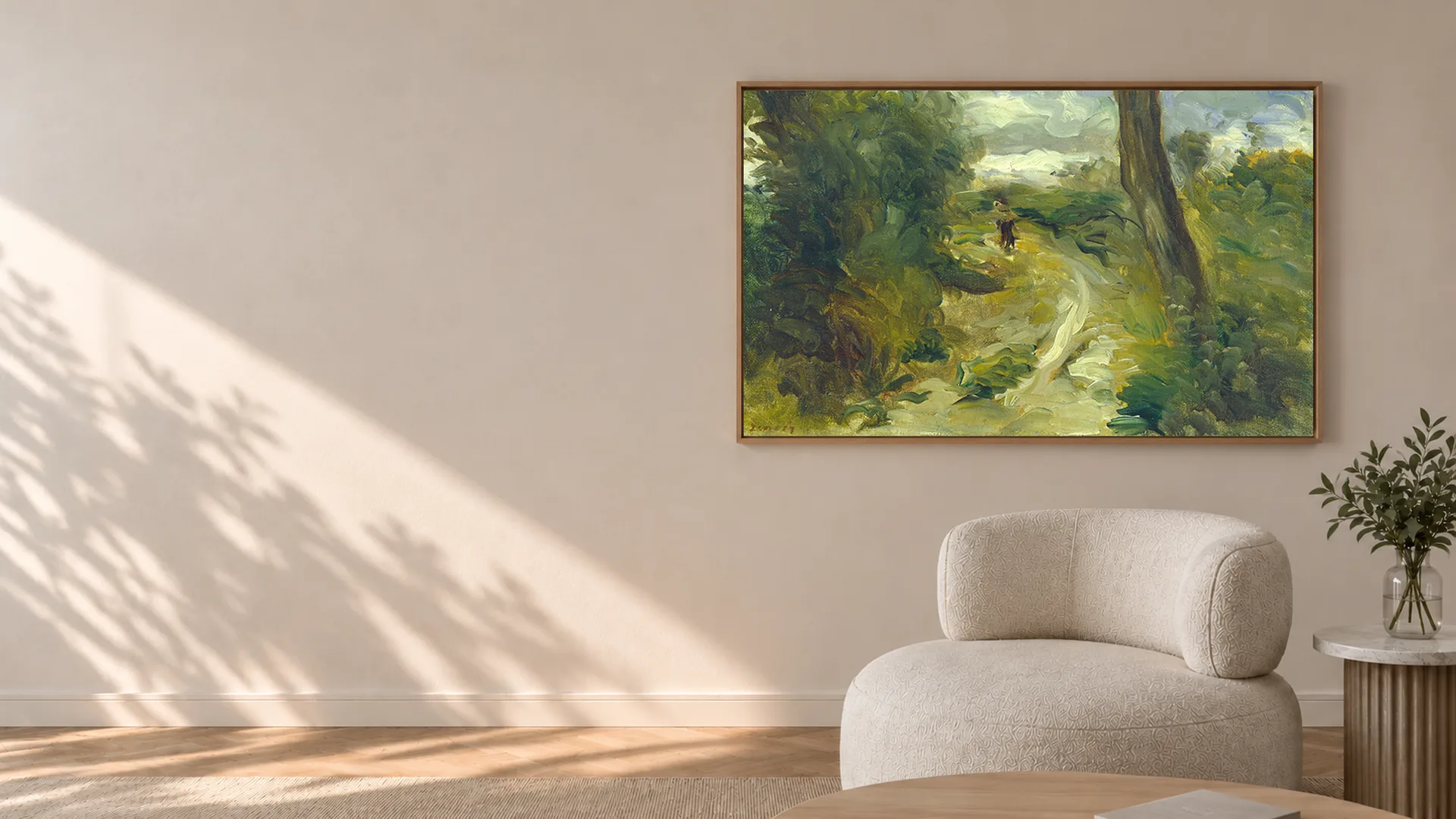 A landscape painting of a path through trees, in a slim oak frame on a warm plaster wall in afternoon light and leaf shadow, beside a boucle armchair and a small olive tree