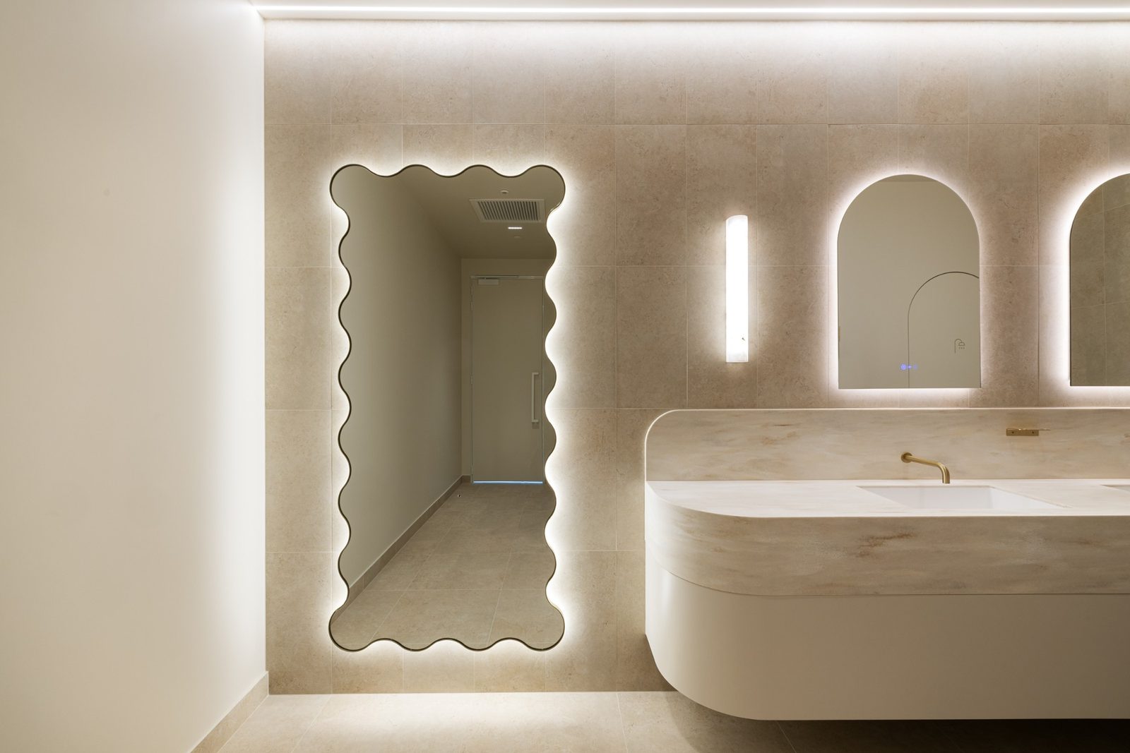 A full-height scalloped mirror, edge-lit with a warm halo, in a pale stone bathroom