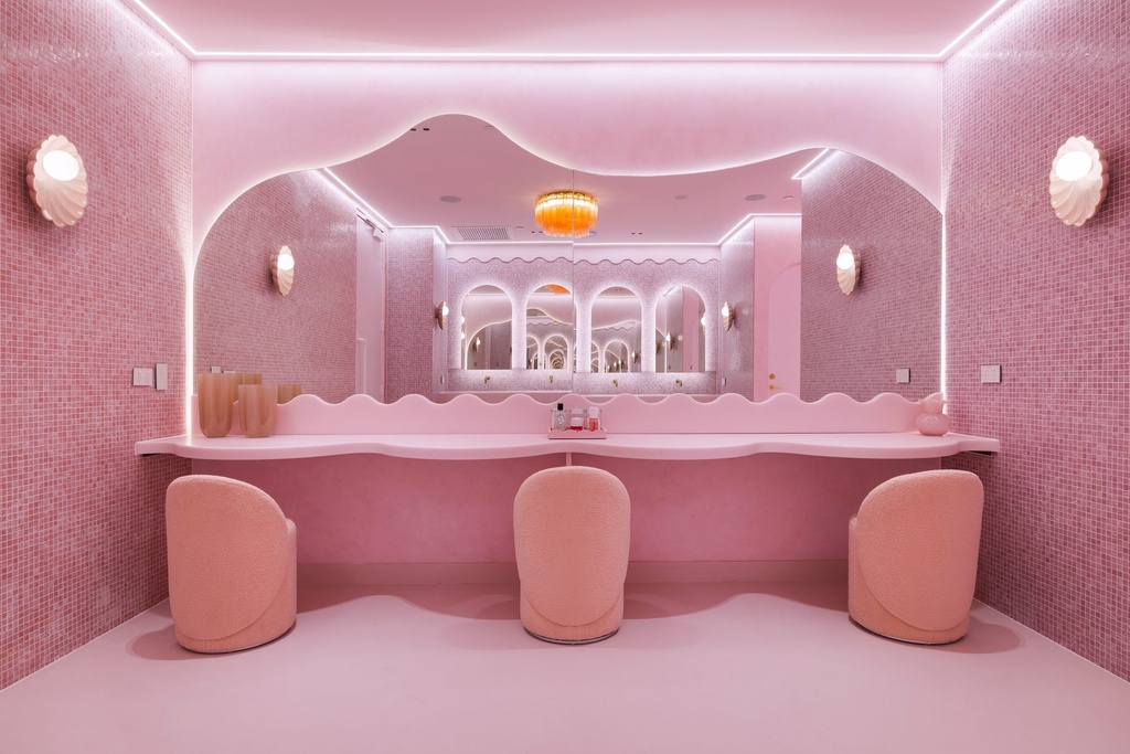 A pink tiled powder room with a wide mirror whose top edge waves, lit from behind, above a curved bench and three pink chairs