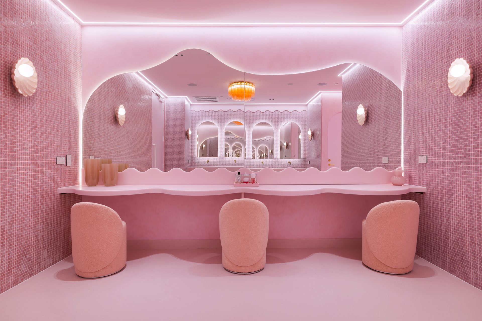 A pink tiled powder room with a wide mirror whose top edge waves, lit from behind, above a curved bench and three pink chairs
