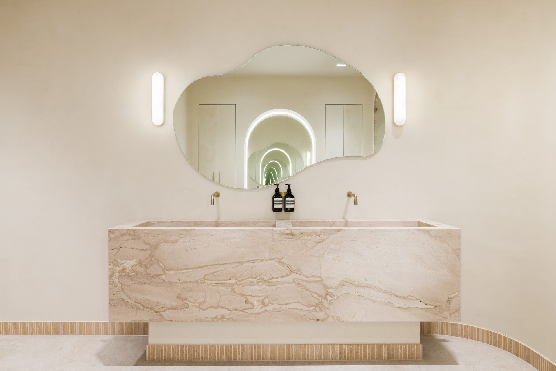 A frameless organic-shaped mirror between two wall lights above a marble trough basin