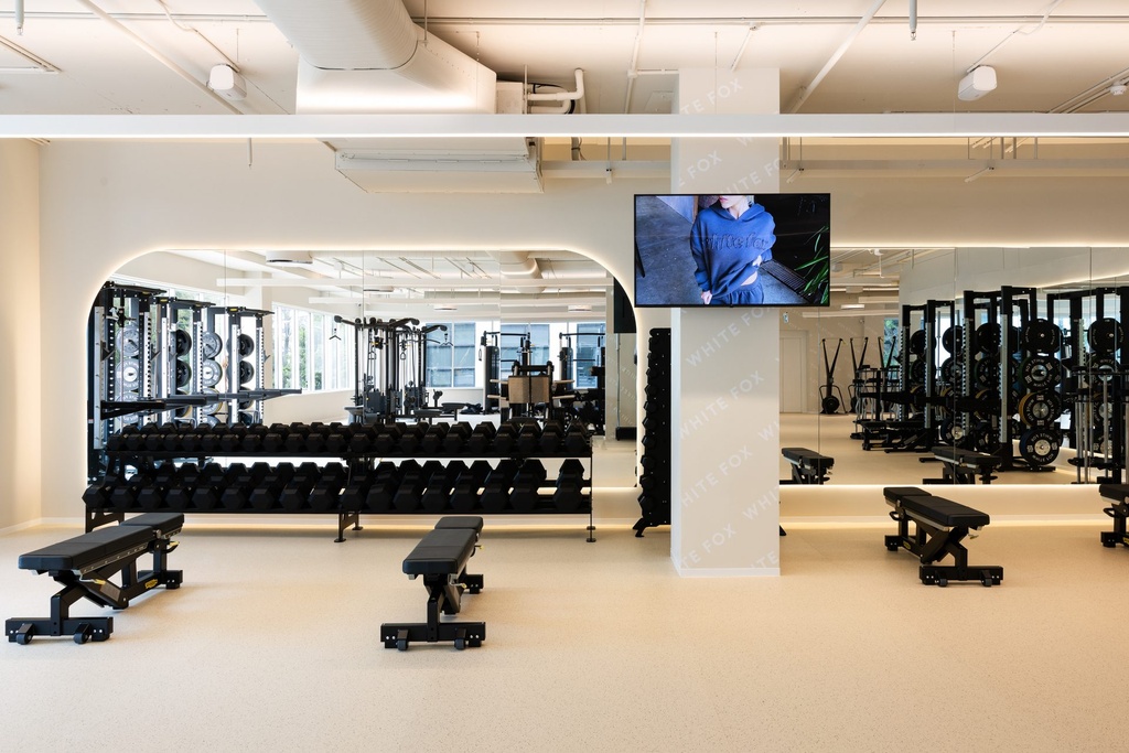 A gym with dumbbell racks and benches in front of a long mirror wall with rounded top corners, lit along its edge