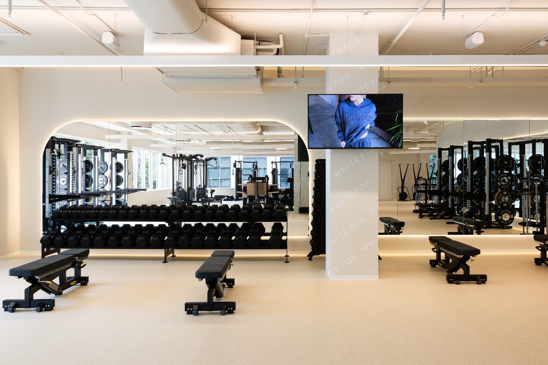 A gym with dumbbell racks and benches in front of a long mirror wall with rounded top corners, lit along its edge
