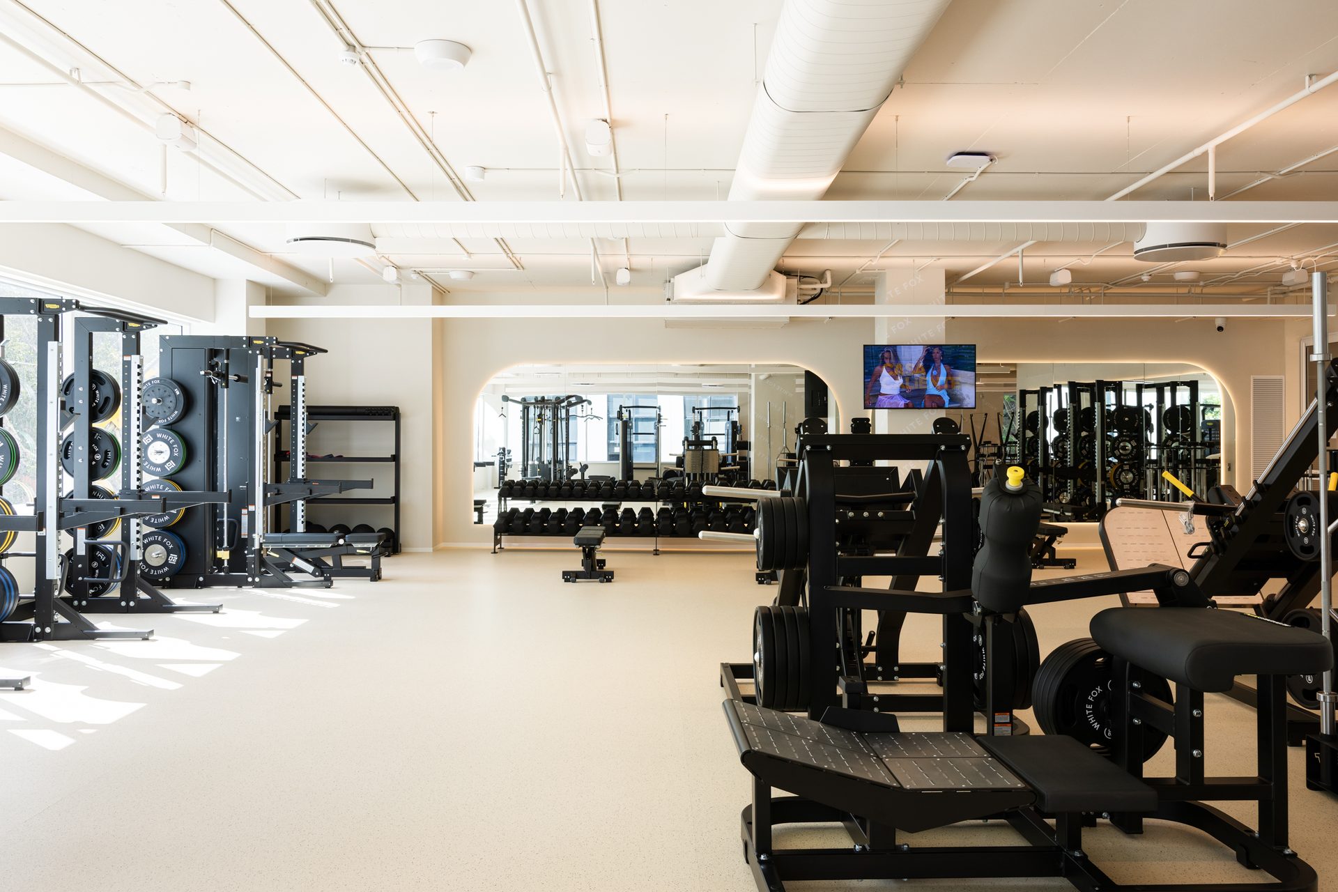 A gym floor with weight racks and machines, and a mirror wall with rounded corners at the far end