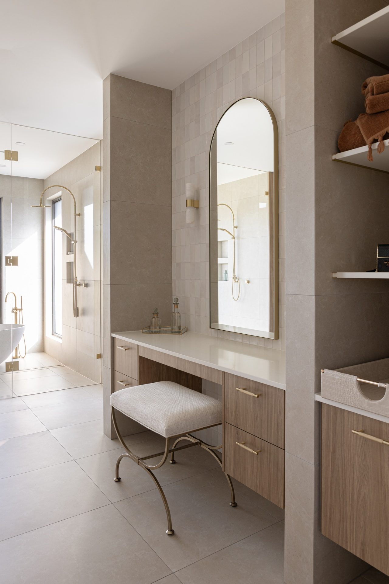 A dressing table with an upholstered stool under a tall arched mirror in a slim brass frame, with the shower beyond