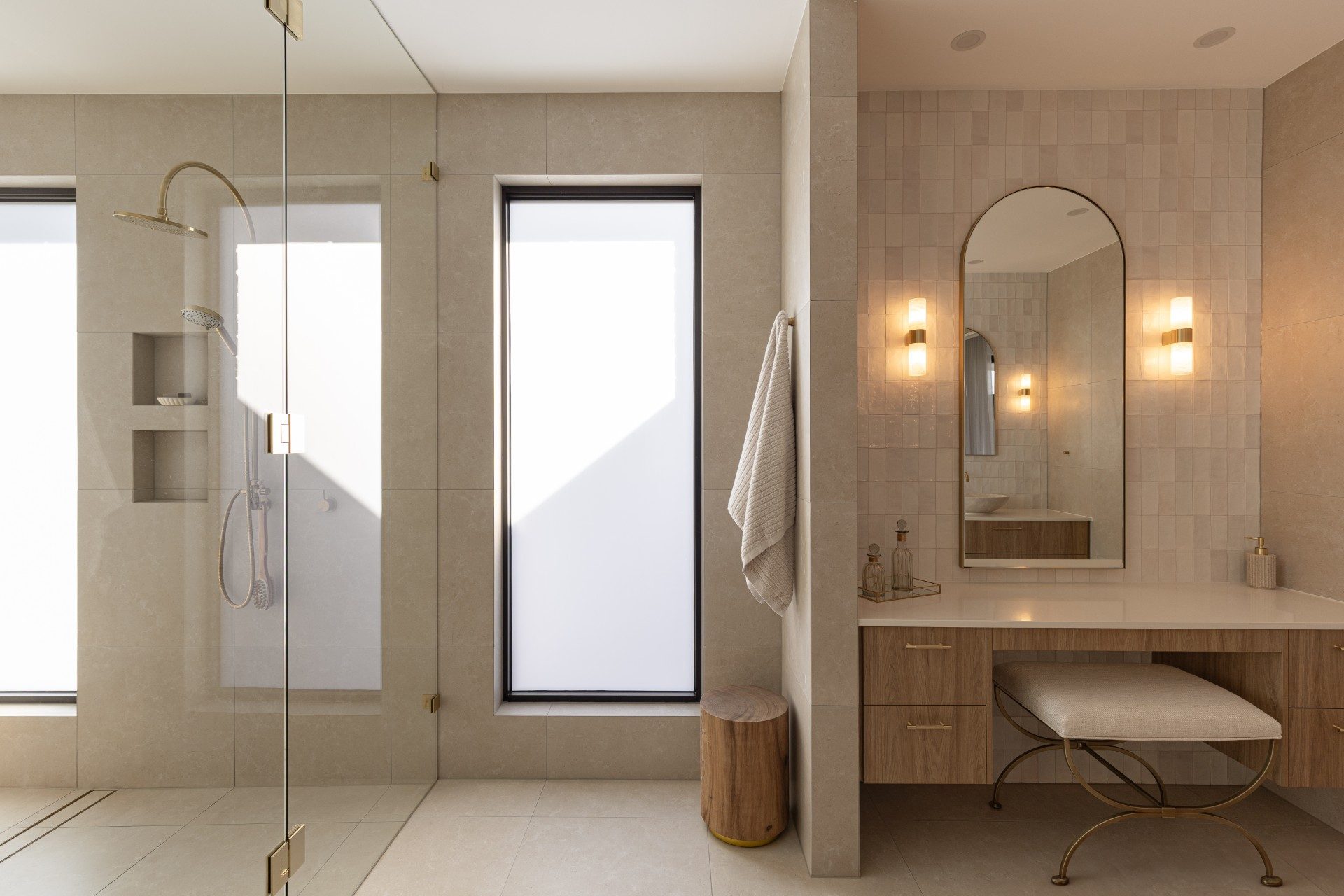 An ensuite with a glass-walled shower on the left and a tall arched mirror in a slim brass frame above a dressing table on the right