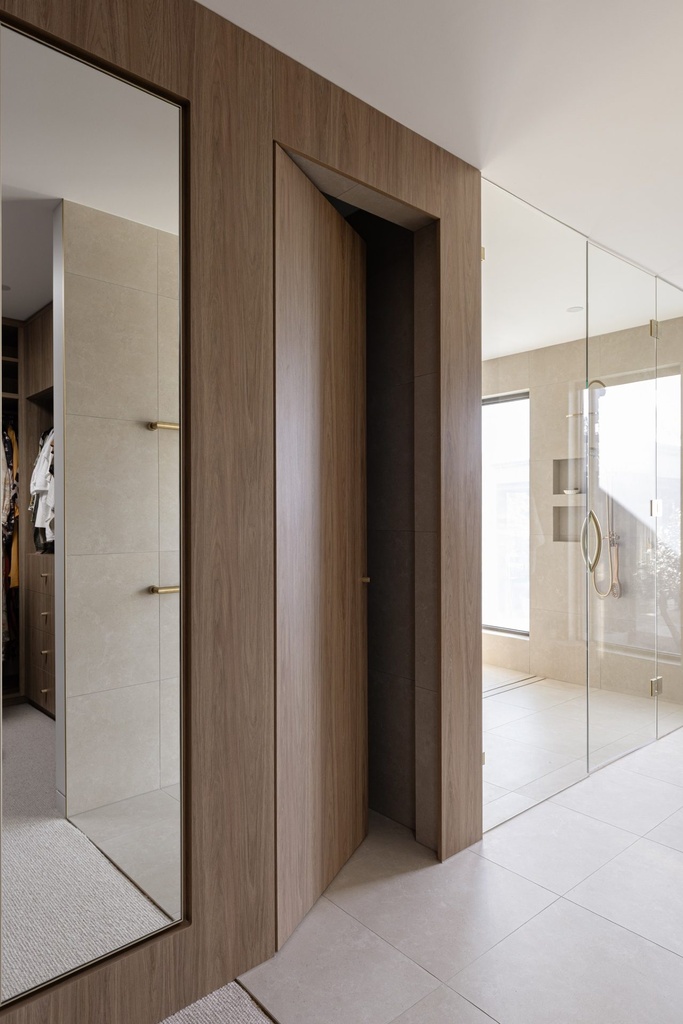 A tall mirror in a slim dark frame on a timber wall beside a hidden door, with the ensuite shower beyond
