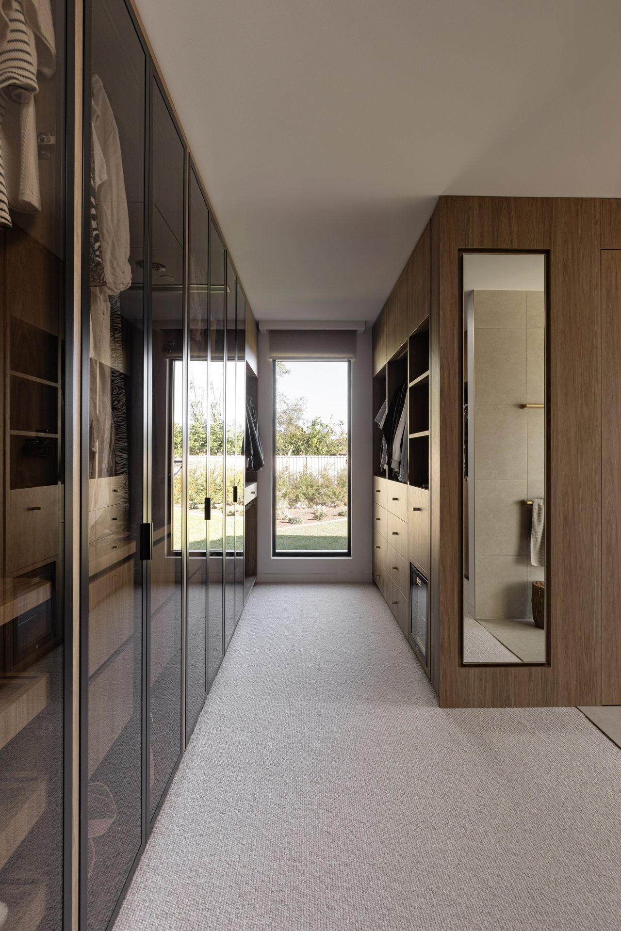 A walk-in robe with glass-fronted wardrobes on the left and a tall framed mirror set into the timber joinery on the right