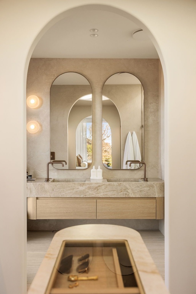 View through an arched doorway to two tall arched mirrors above a stone double vanity, with a stone-topped island in the foreground