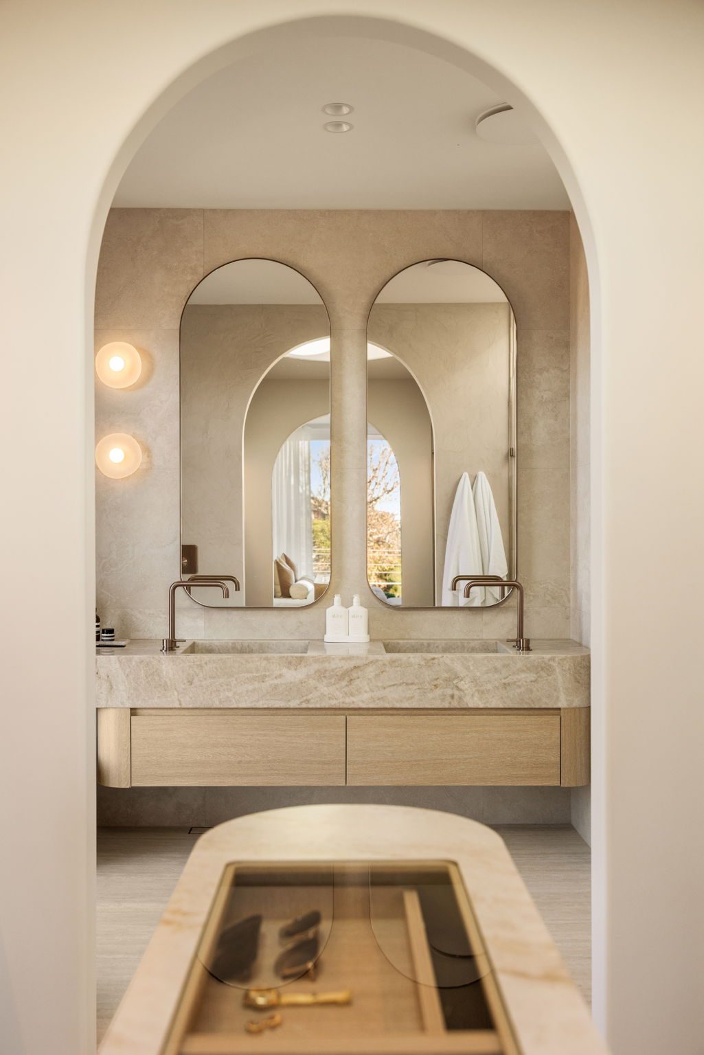View through an arched doorway to two tall arched mirrors above a stone double vanity, with a stone-topped island in the foreground