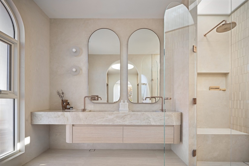 Two tall arched mirrors in slim frames above a floating stone vanity with two copper taps, with the shower to the right