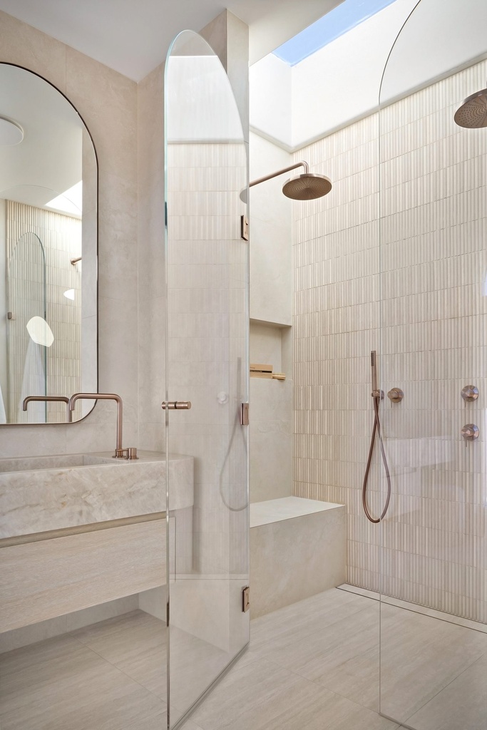 A tall arched mirror above a stone vanity next to a glass shower screen with an arched top, under a skylight