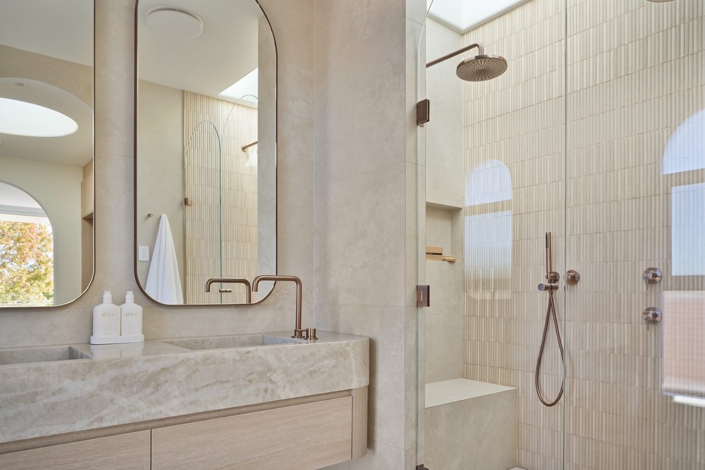 Two tall mirrors with rounded corners above a stone vanity, beside a glass shower with vertical tiles and copper fittings