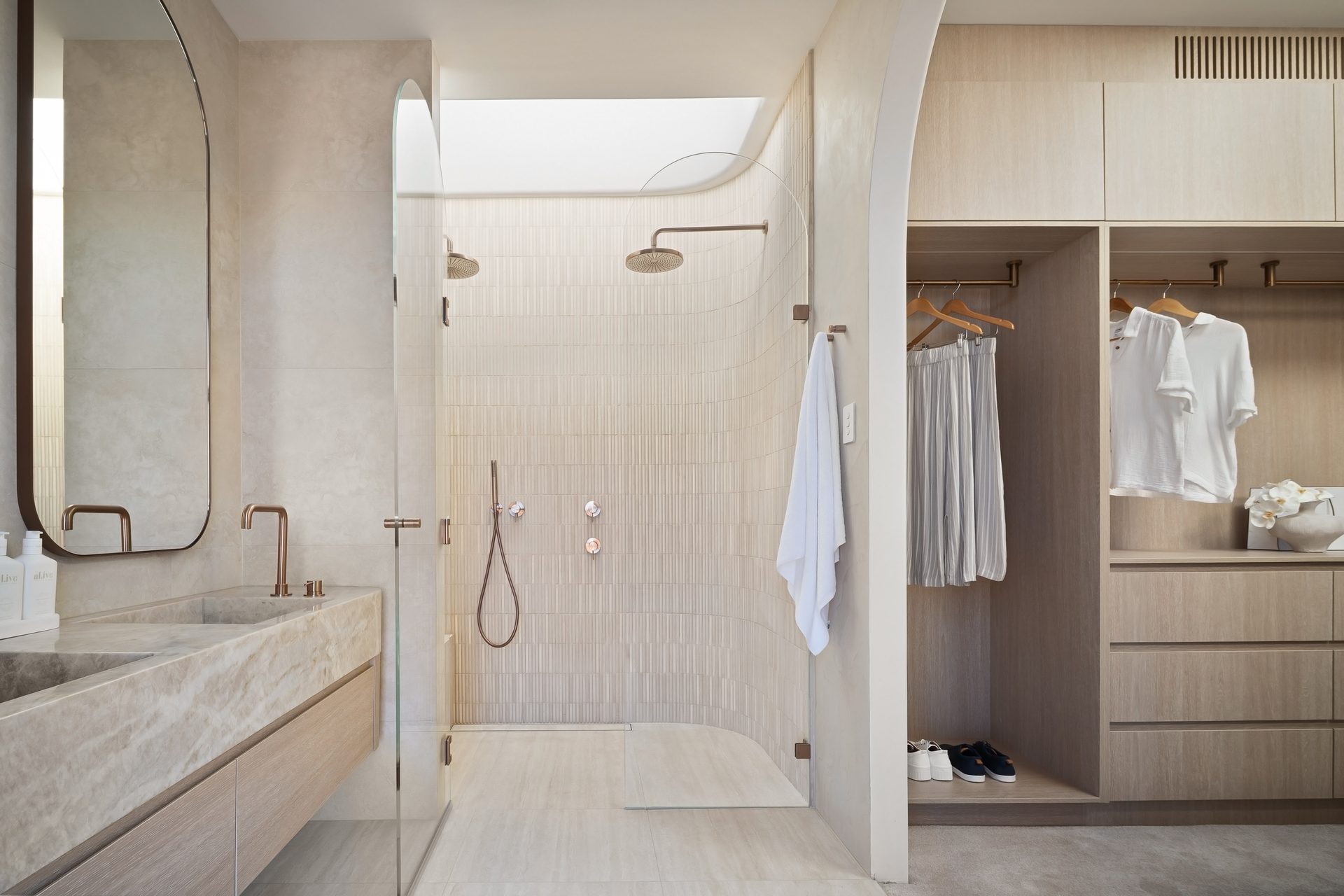 An ensuite with a mirror over the stone vanity, a curved shower under a skylight and an open dressing room to the right