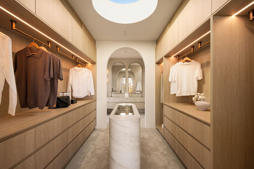 A dressing room with timber wardrobes, lit hanging rails and a stone island, looking through an arch to the twin arched mirrors