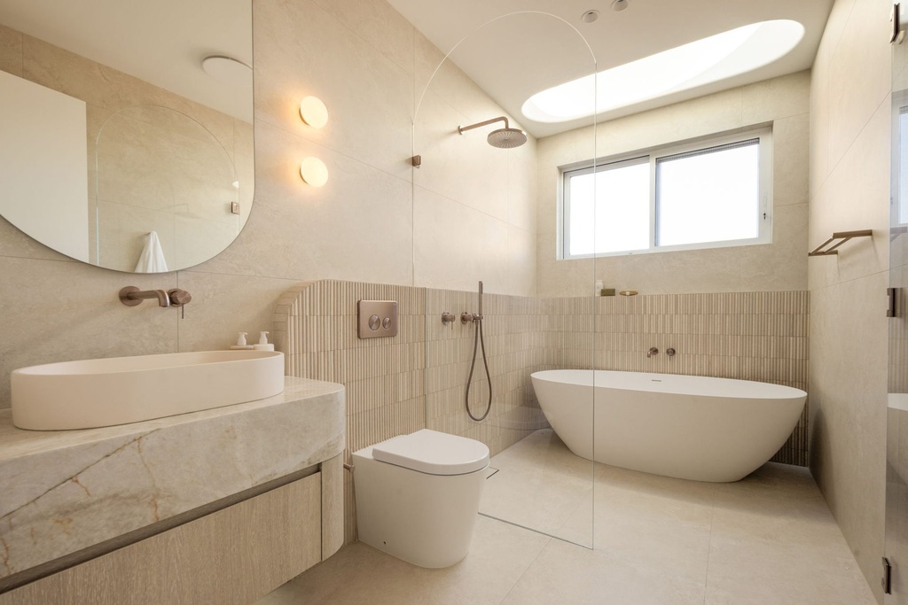 A bathroom with a large frameless mirror with a rounded side above an oval basin on a stone bench, and a freestanding bath behind a glass screen