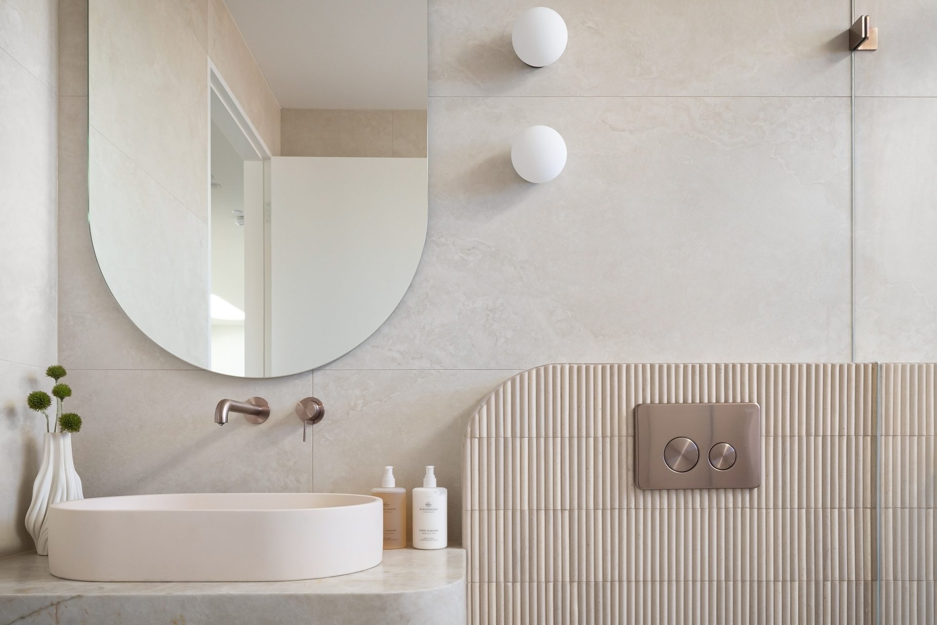 A frameless mirror with one straight and one rounded side above an oval basin on stone, beside fluted tiles and two globe wall lights