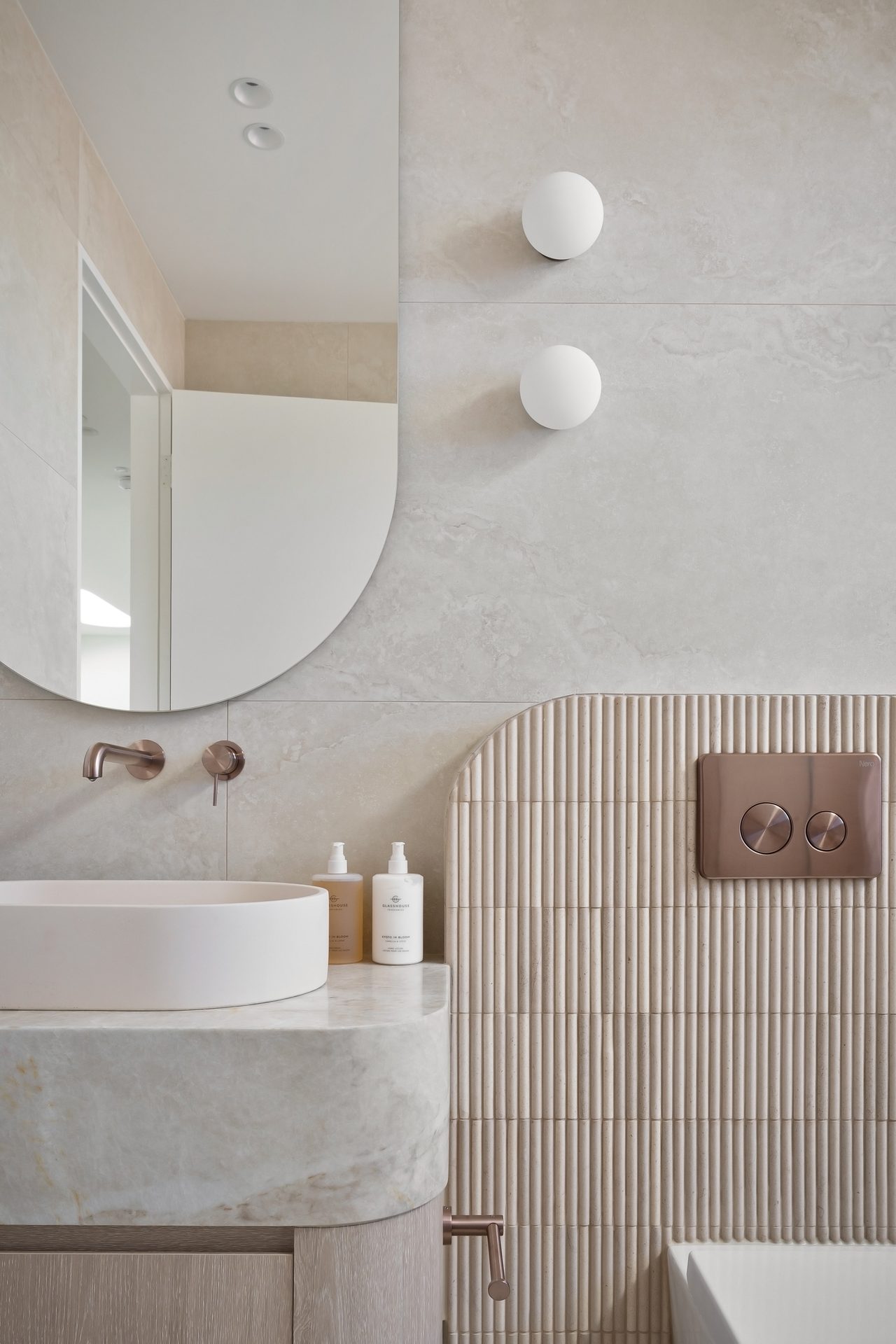Close view of a frameless mirror with a rounded edge above a basin, with globe wall lights and fluted tiles