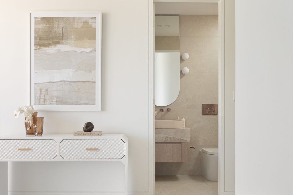 A white console under a framed abstract artwork, beside an open door to a bathroom with a round-edged mirror