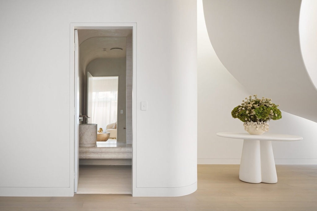 A white hallway with a curved wall and a round side table, and an open door showing a curved-top mirror in the room beyond