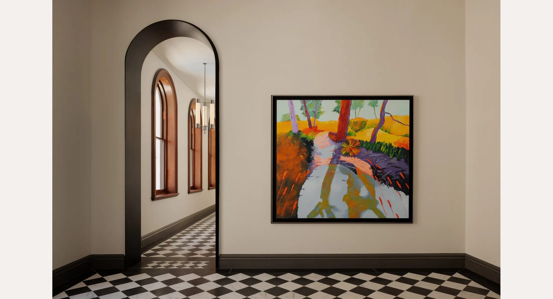 A square landscape painting of a red tree trunk beside a path, in a dark frame, next to an arched doorway over a chequered floor