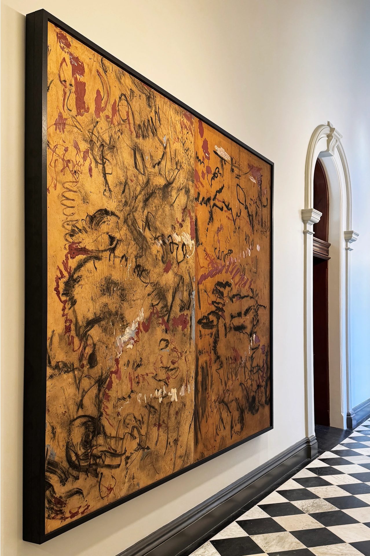 A large ochre abstract painting with black and red marks in a dark frame, in a hallway with an arched doorway