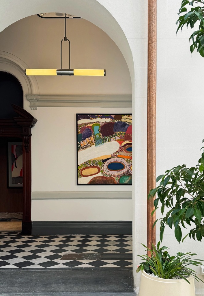 A large colourful painting of rounded landforms and waterholes in a dark frame, seen through an arch from a light-filled atrium