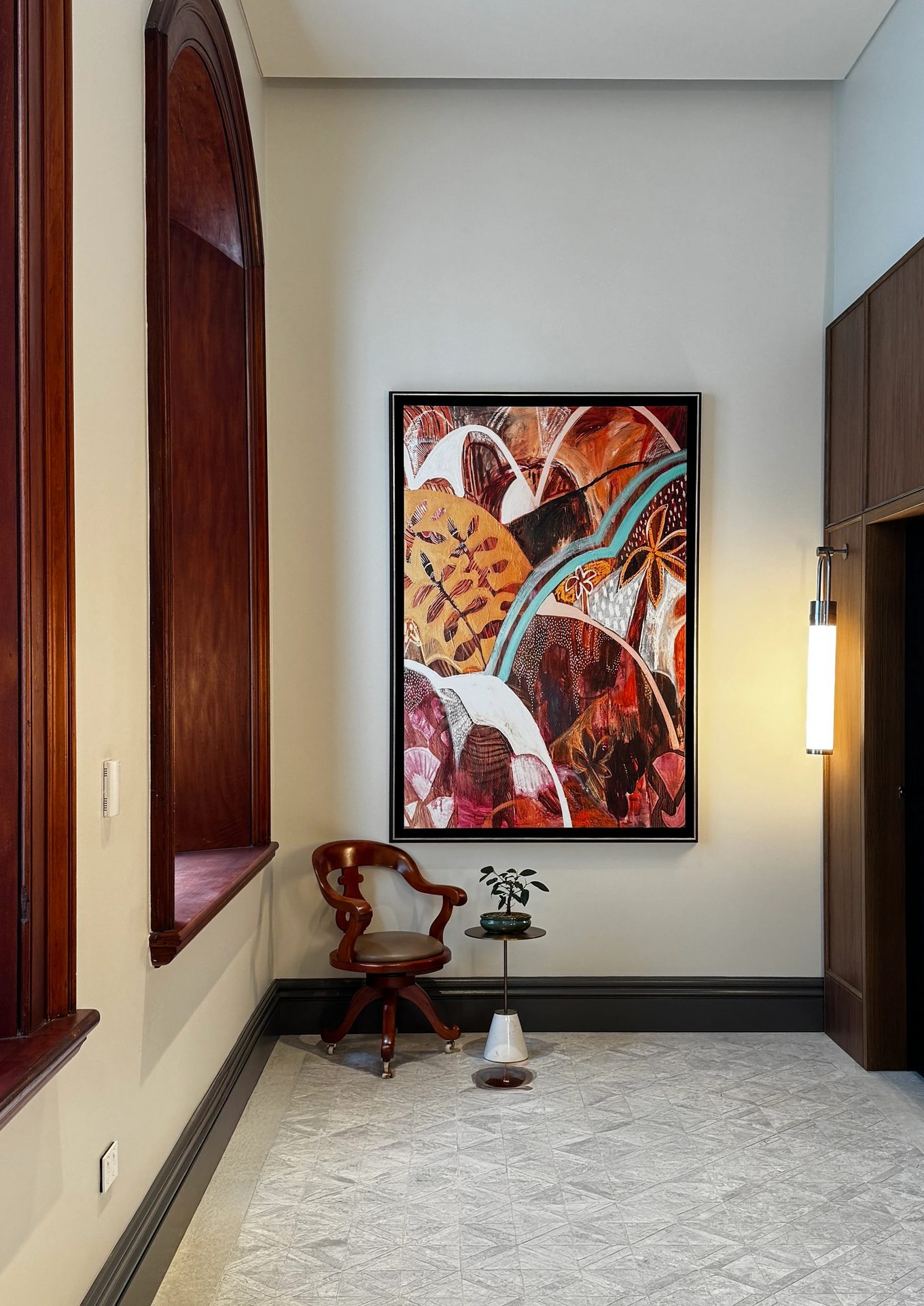 A tall painting of red, white and teal arcs and leaves in a dark frame, in a high room with arched timber windows and a leather chair