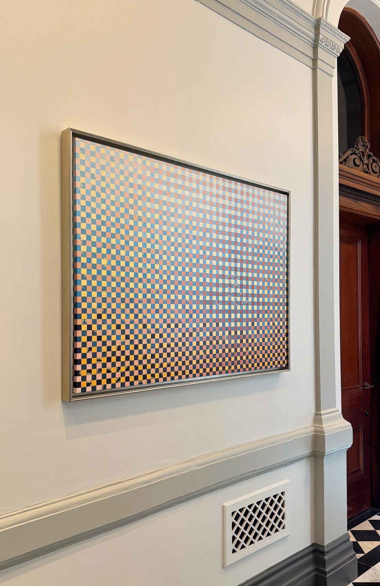 A wide painting of a fine multicoloured grid in a pale metal frame, on a wall with heritage mouldings