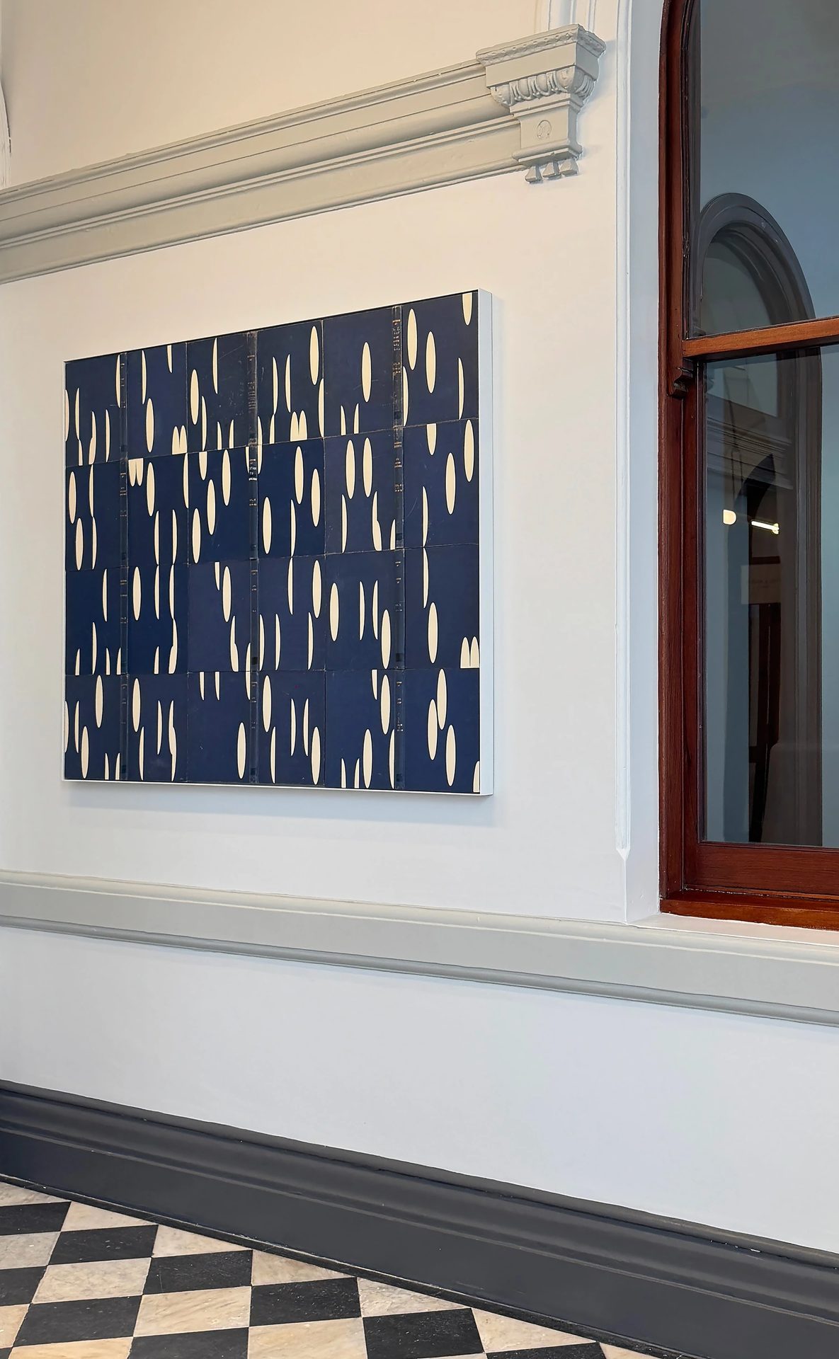 A large panel work of cream ellipses on deep blue in a white frame, on a heritage wall beside a timber-framed window