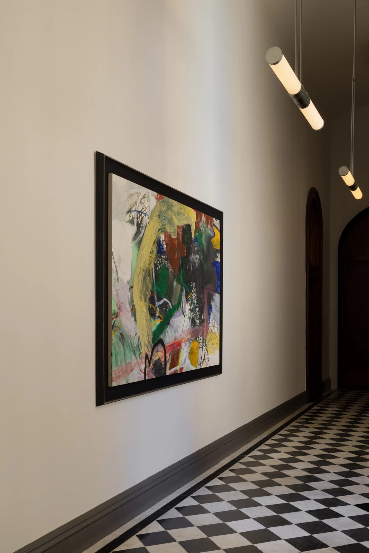 A large gestural abstract painting in yellow, green and black in a dark frame, along a corridor with pendant lights