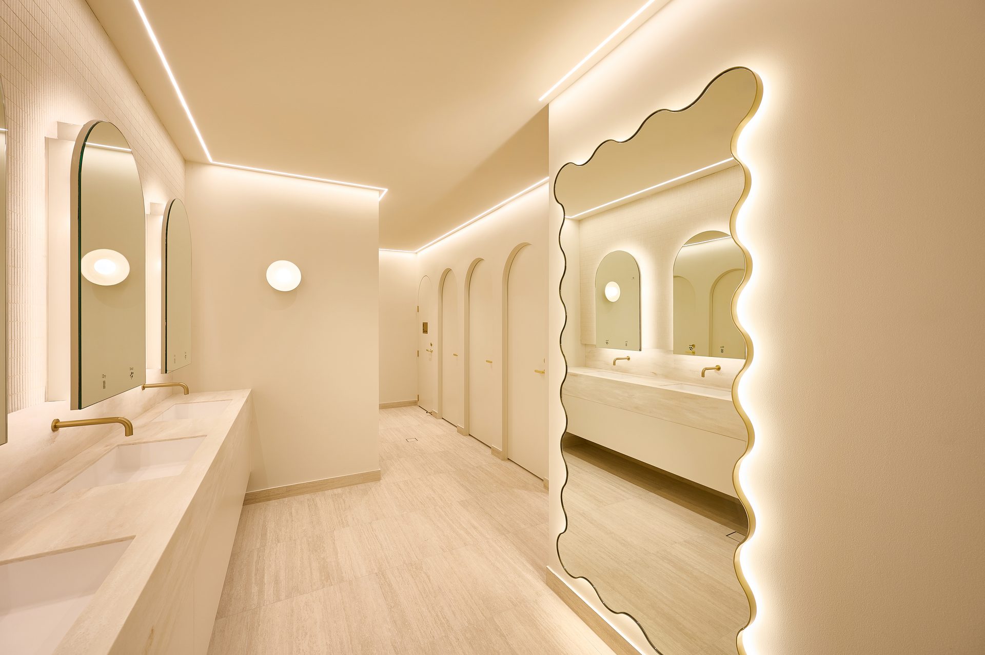 A long amenities room with a tall wavy-edged backlit mirror on the right and arched mirrors over a stone vanity on the left