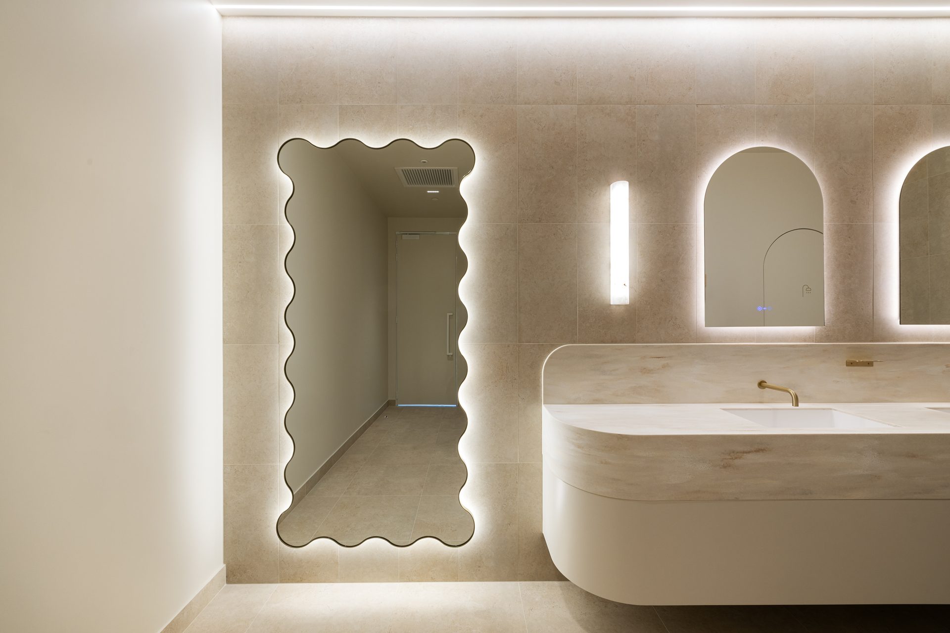 A tall mirror with a wavy edge and a warm halo of light on a stone-tiled wall, next to arched backlit mirrors above a curved stone vanity