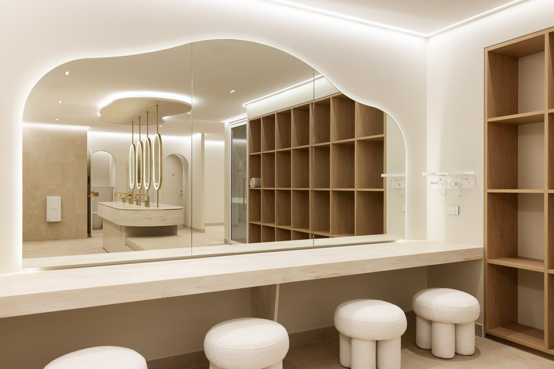A wide mirror with a flowing top edge, lit from behind, above a long dressing bench with round upholstered stools