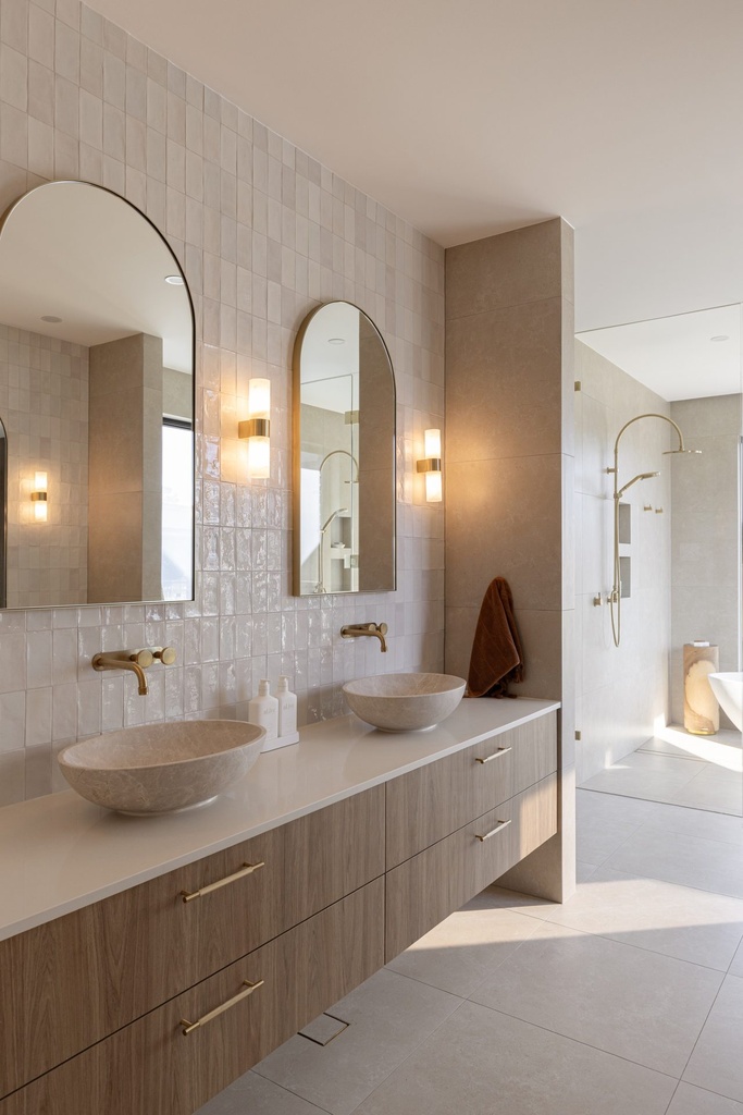A double vanity with two stone vessel basins under two arched mirrors in slim brass frames, between glass wall lights