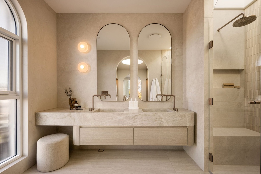 An ensuite with two tall arched mirrors in slim frames above a floating stone double vanity, beside an arched window and a glass shower